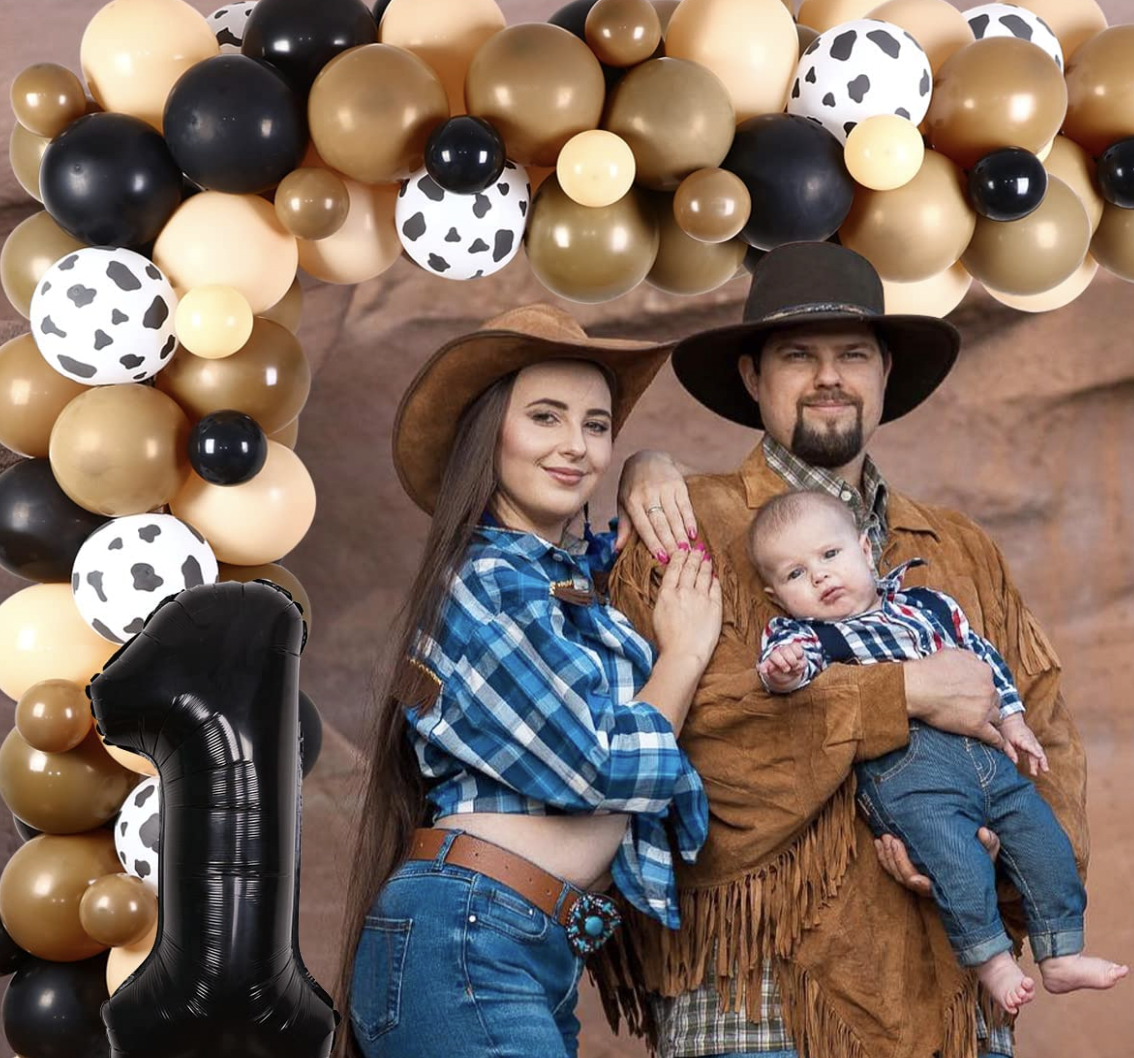A woman in American western style clothing leans on a man dressed in western clothes while he holds a child in overalls. The family stands in front of a balloon arch with a number one balloon.