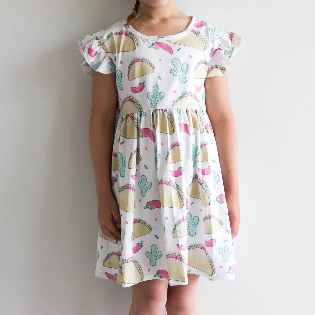 Child wearing white dress with pattern of tacos, chili peppers, and cacti.