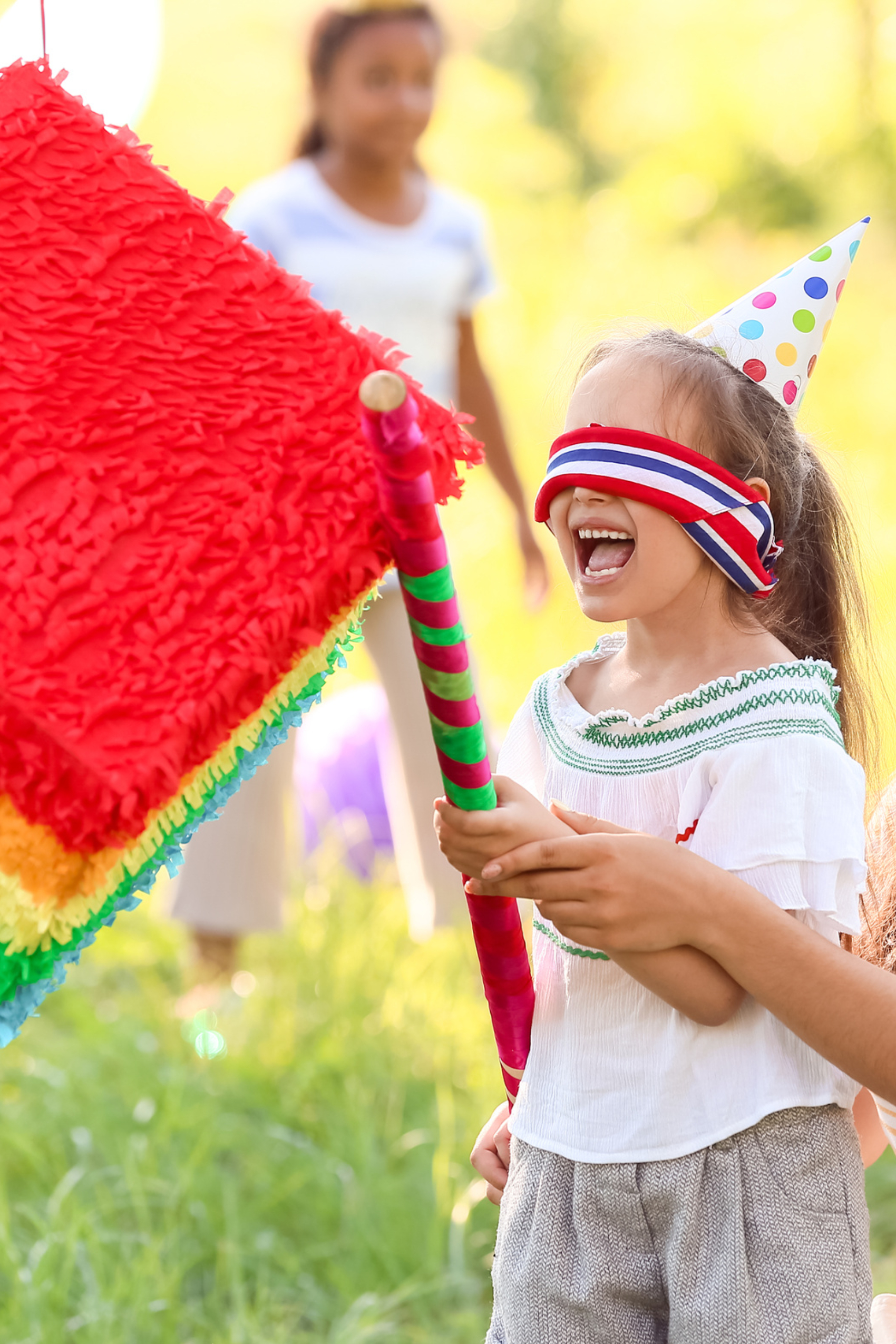 A young girl wearing a party hat and blindfolded is guided to where the pinata is hanging.